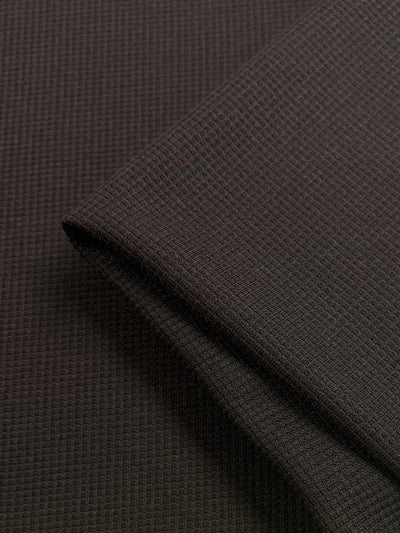 A close-up view of a dark brown fabric with a textured, waffle weave pattern. The Waffle Knit - Brown - 170cm by Super Cheap Fabrics is neatly folded, showcasing its fine, grid-like texture and three-dimensional effect.