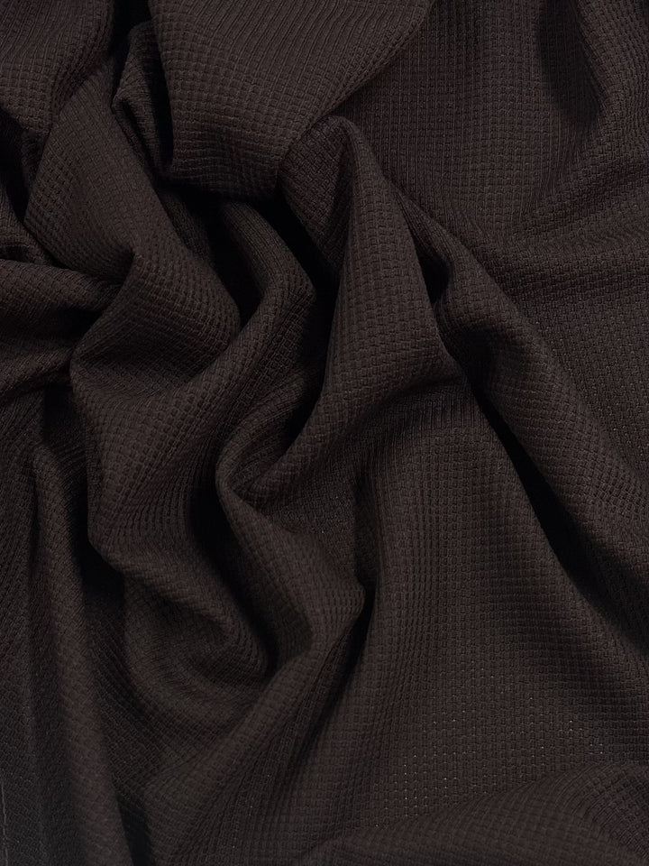 The Waffle Knit - Brown - 170cm fabric by Super Cheap Fabrics draped in soft folds, creating an intricate pattern of ridges and valleys with a three-dimensional effect. The texture appears cozy and inviting, suggesting a warm and comfortable material.