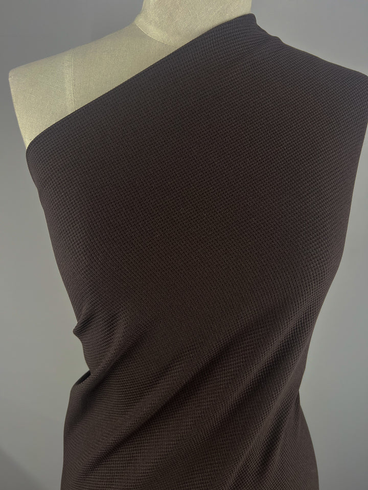 A textured, one-shoulder Waffle Knit - Brown - 170cm by Super Cheap Fabrics draped over a bust form against a plain gray background. The slightly stretchy material boasts a subtle honeycomb pattern, creating a sleek, tailored look with a gentle three-dimensional effect.