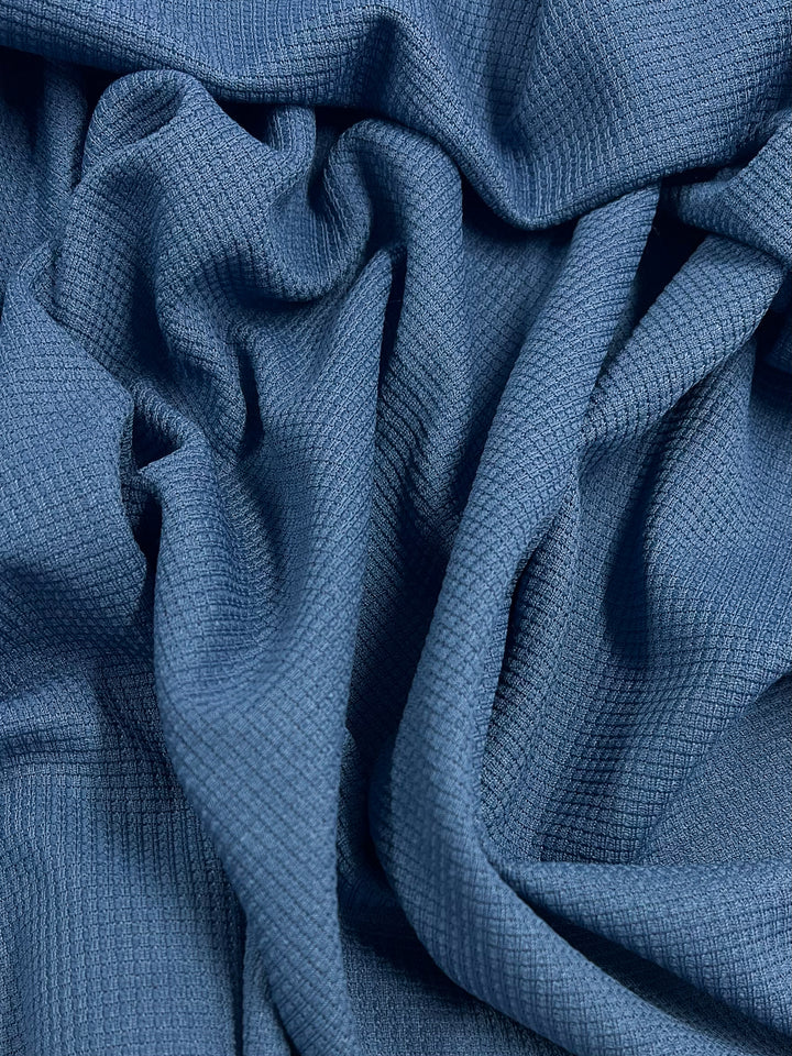 A close-up view of Super Cheap Fabrics' Waffle Knit - Stellar - 170cm in blue, showcasing its textured, waffle-knit pattern. The soft and flexible fabric forms intricate folds and shadows, giving it a three-dimensional appearance.