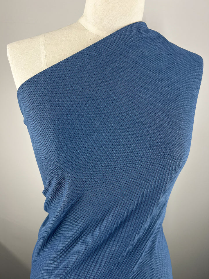 A mannequin adorned in Super Cheap Fabrics' Waffle Knit - Stellar - 170cm, featuring a textured blue honeycomb pattern that gracefully drapes around the shoulders. The plain, neutral background accentuates the fabric's vibrant color and intricate three-dimensional design.