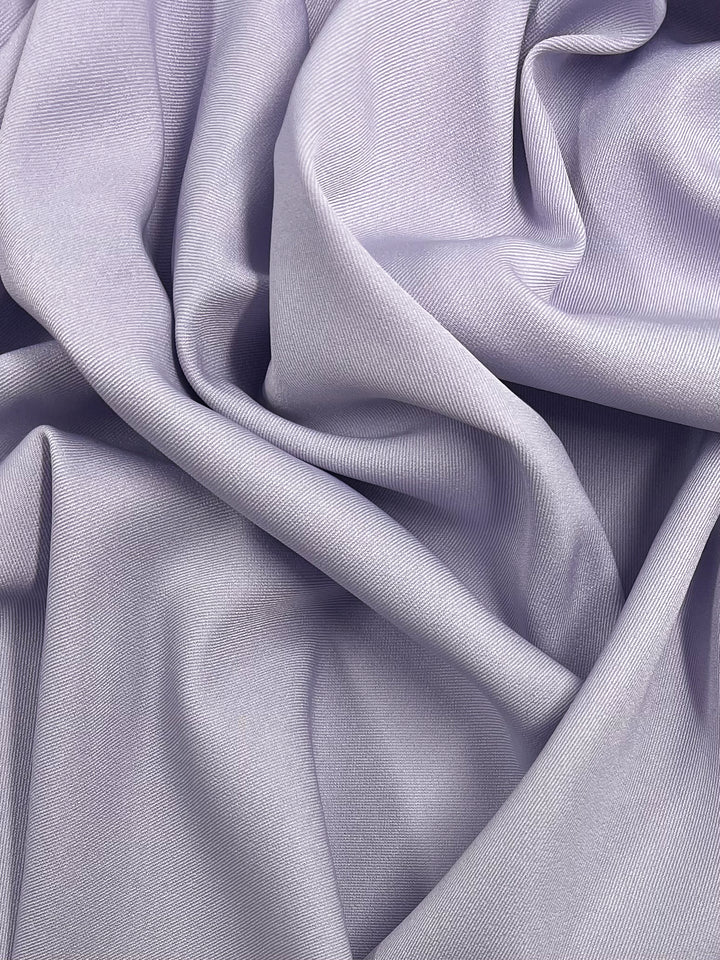 A detailed image of the Twill Suiting - Lavender Blue from Super Cheap Fabrics, showcasing its soft, light purple hue with a smooth, slightly shiny texture. The versatile fabric is artistically draped and folded in multiple overlapping layers, creating gentle creases and shadows that add depth to the material.