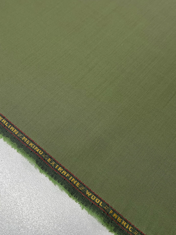 Close-up of a green fabric with a black and yellow selvedge edge. The text on the selvedge reads "Italian Merino Extrafine Wool Fabric" in all caps. The luxurious texture of the Merino Wool Suiting - Hunter Green - 155cm by Super Cheap Fabrics is evident as it lies on a flat white surface, promising both elegance and breathability.