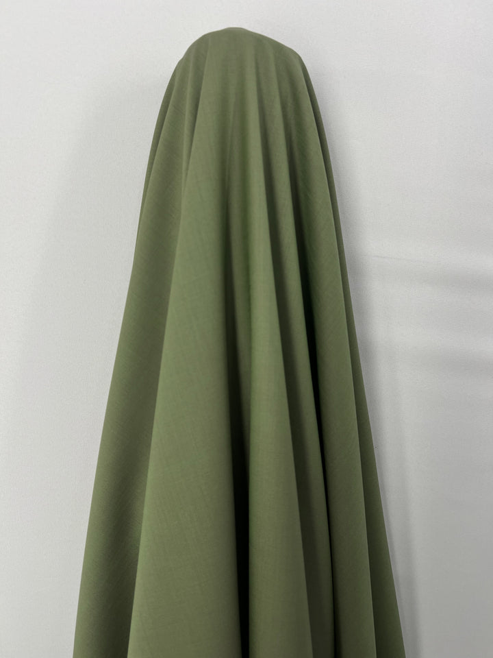 A piece of Merino Wool Suiting - Hunter Green - 155cm from Super Cheap Fabrics, with a luxurious texture, draped over a hidden object against a plain white wall. The material hangs smoothly, creating soft folds and shadows.