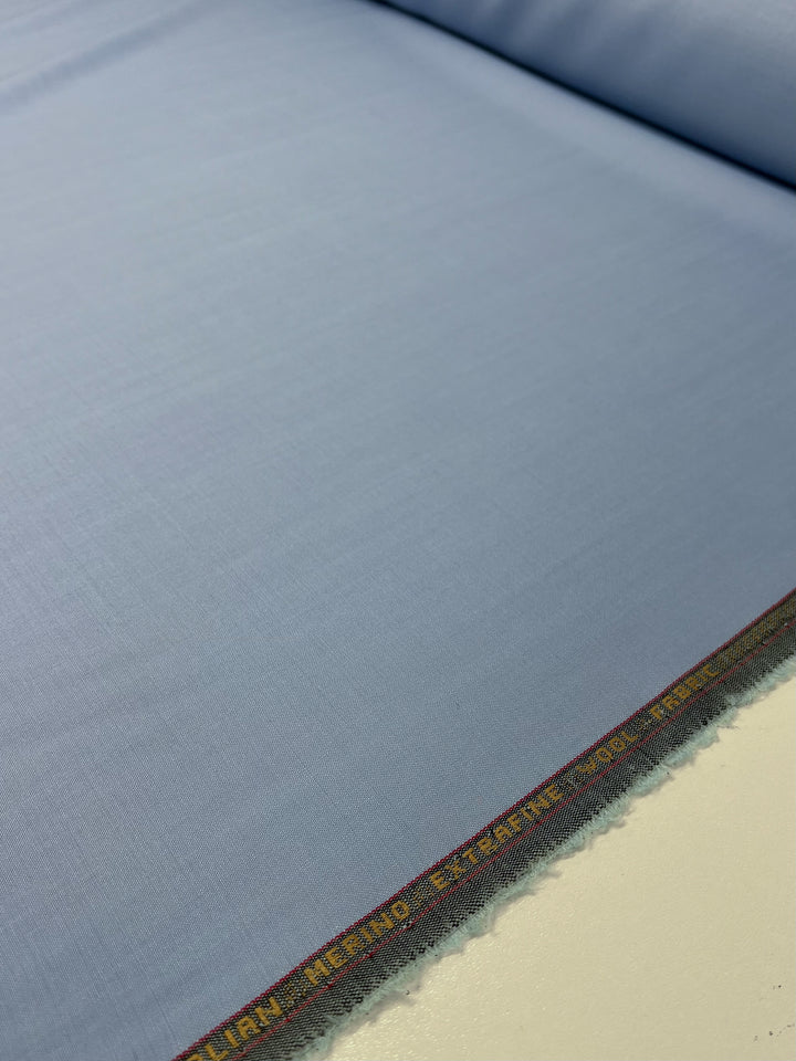 A close-up image of a roll of light blue **Super Cheap Fabrics Merino Wool Suiting - Serenity - 155cm**. The luxurious texture's edge has a multicolored stitching detail, and there is text in yellow thread running along the border. The fabric appears smooth and is laid out flat.