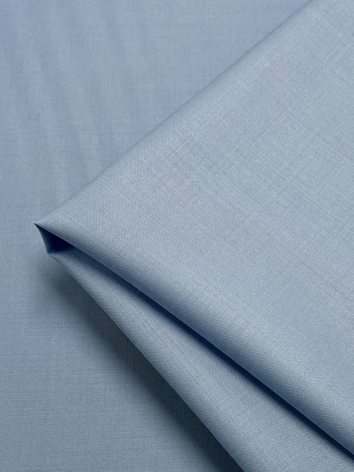 A close-up shot of neatly folded light blue fabric with luxurious texture. The smooth fabric, featuring a subtle weave pattern, boasts a clean and polished appearance. The image focuses on the folds and the material's fine details, revealing its moisture-wicking properties. This is the Merino Wool Suiting - Serenity - 155cm by Super Cheap Fabrics.
