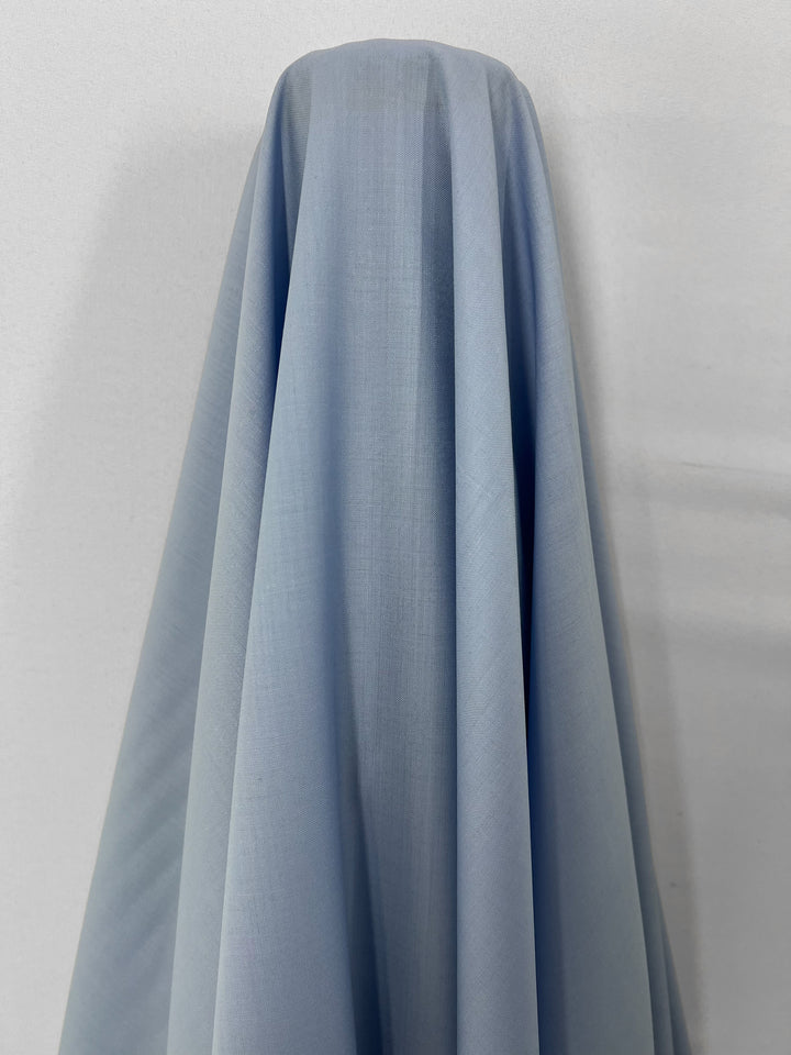 A light blue fabric draped over an object, creating soft folds and shadows. The background is plain and off-white, highlighting the luxurious texture and smooth, semi-sheer nature of the cloth. The product in question is the Merino Wool Suiting - Serenity - 155cm by Super Cheap Fabrics.