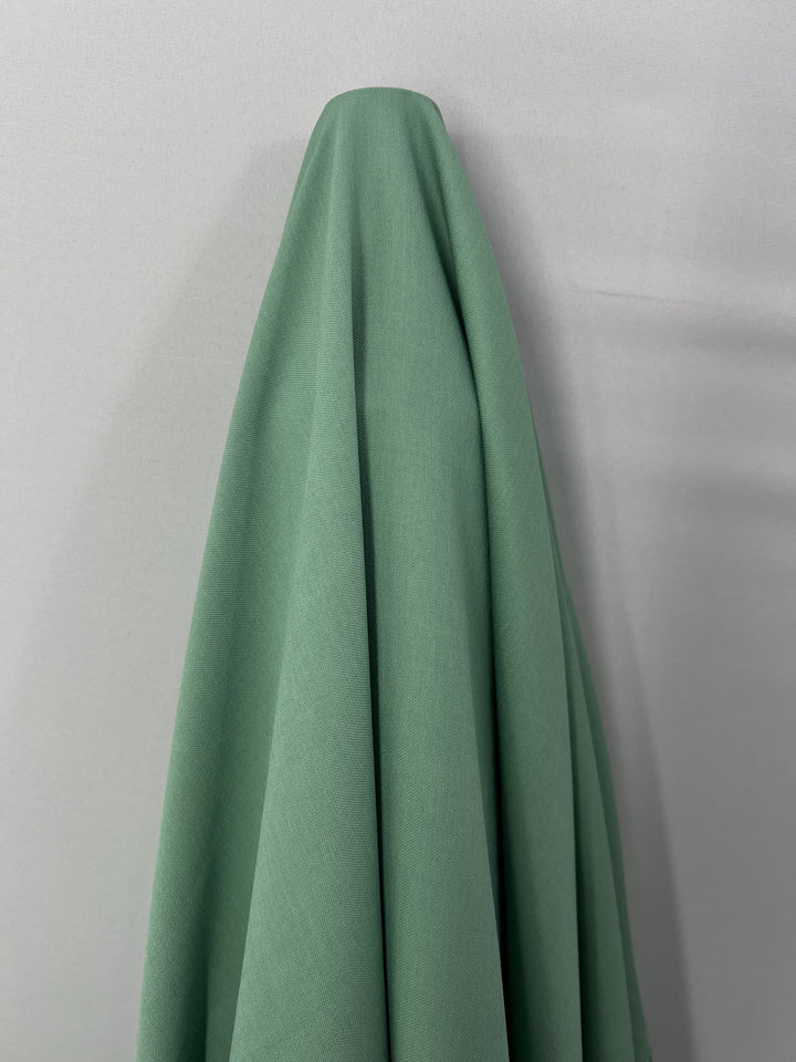 A piece of Bamboo Suiting - Hemlock - 140cm by Super Cheap Fabrics hangs against a plain, light-colored wall. The lightweight fabric forms soft folds and drapes down smoothly, creating a minimalist and serene visual.