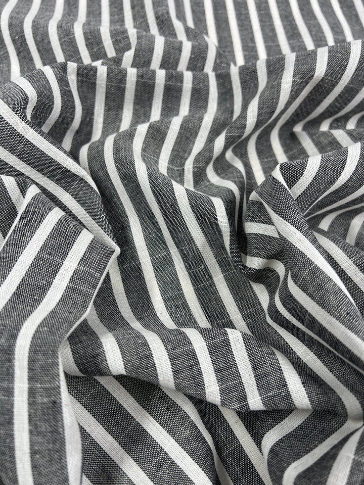 Close-up of crumpled fabric with a pattern of alternating light and dark vertical stripes. The Super Cheap Fabrics' Linen Cotton - Dark Grey Stripe - 145cm boasts a soft, textured appearance, with shadows and folds creating depth. The durable fabric's evenly spaced stripes are parallel, highlighting the use of natural fibers.