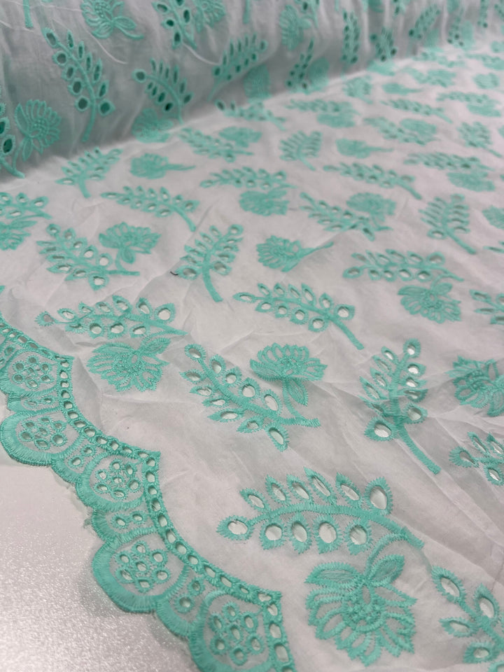 A close-up of a fabric with intricate turquoise floral embroidery on a sheer white background. The edge, reminiscent of Broderie Anglaise, showcases a scalloped pattern with detailed floral designs and small circular cutouts, enhancing the fabric's delicate beauty. This is none other than the Broderie Anglaise - Aqualotus - 125cm by Super Cheap Fabrics.