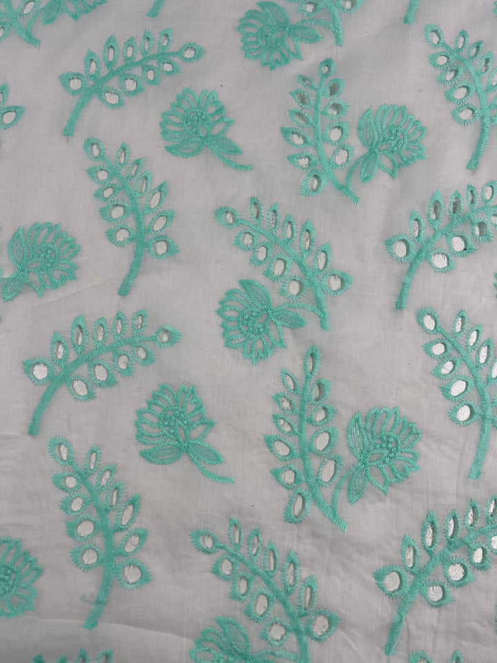 A white polyester fabric is decorated with an intricate turquoise floral and leafy design. The Broderie Anglaise - Aqualotus - 125cm by Super Cheap Fabrics features circular cutout details within the leaf motifs, creating a detailed and delicate pattern across the material.