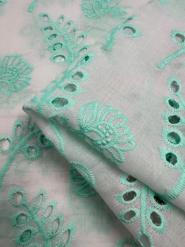 A piece of *Broderie Anglaise - Aqualotus - 125cm* by *Super Cheap Fabrics* featuring intricate teal floral and leaf embroidery, echoing a Broderie Anglaise style. The fabric is folded neatly, showcasing delicate cutout designs and detailed stitching. Soft and lightweight, it’s ideal for clothing or home decor.