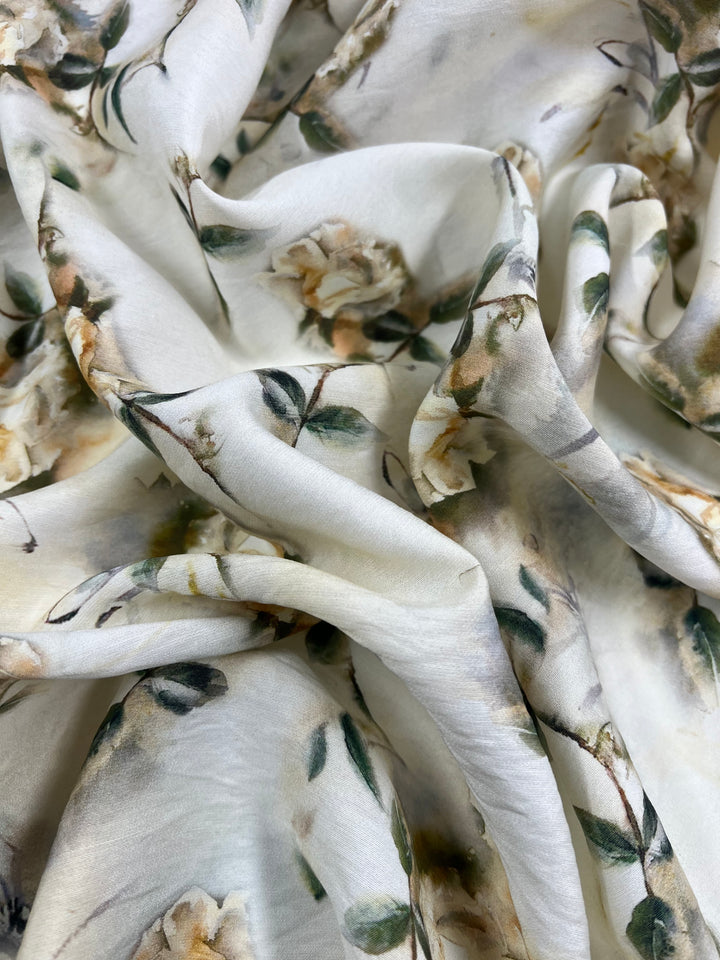 A close-up of soft, white fabric with a floral pattern featuring light beige flowers and green leaves, perfect for children's clothing. The Designer Cotton - Painted Roses - 145cm from Super Cheap Fabrics is gently draped, creating natural folds and shadows that highlight its smooth and delicate texture.