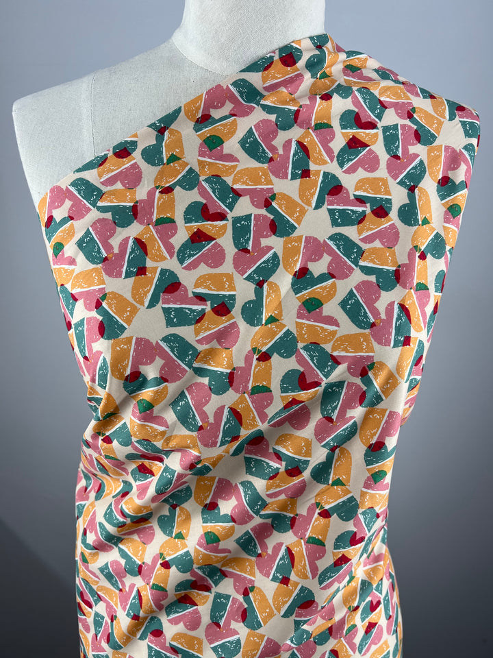 A draped mannequin is adorned with Super Cheap Fabrics' Printed Cotton - Fruity Candy Hearts - 148cm, featuring an abstract pattern of various shapes in shades of pink, orange, green, yellow, and white, set against a light background. The fabric's vibrant and playful design is showcased in the image.