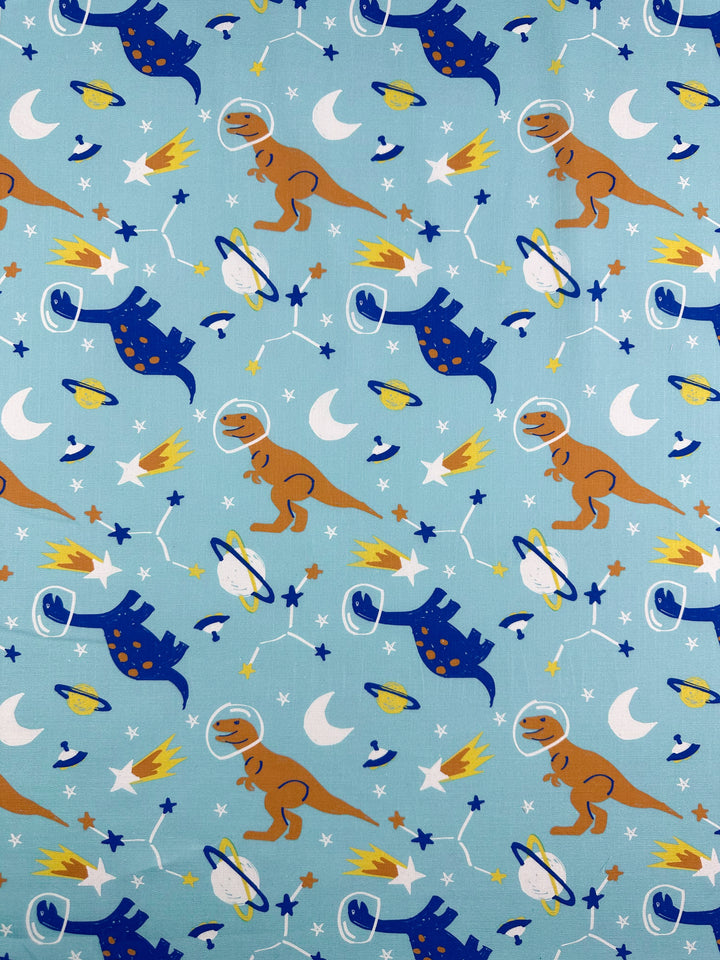 The "Printed Linen - Dino Space - 145cm" by Super Cheap Fabrics features cartoon dinosaurs in helmets, planets, rockets, stars, and moons on a light blue background. The playful orange and blue design is ideal for home decor or imaginative garment creations.