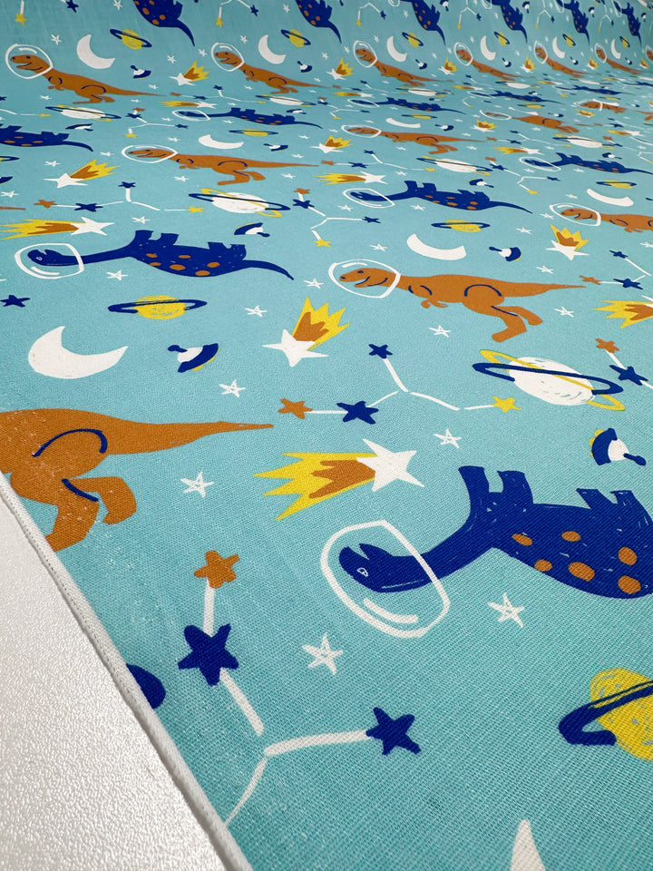 Super Cheap Fabrics' Printed Linen - Dino Space - 145cm features cartoon dinosaurs in space suits floating among stars, planets, and comets on a light blue background. Ideal for garments or home decor, with vibrant blue, orange, and yellow dinosaurs adding a playful touch.