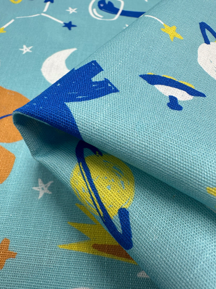 Close-up of Super Cheap Fabrics' Printed Linen - Dino Space (145cm), a blue linen with a vibrant space-themed design featuring planets, rockets, stars, and moons. Ideal for home decor or garments, the slightly folded fabric highlights texture and pattern variations.