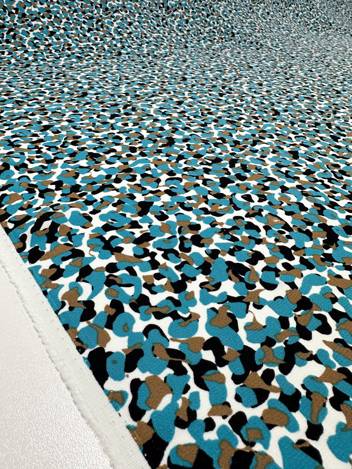 A close-up of the Viscose Crepe - Stoney by Super Cheap Fabrics showcases its luxurious texture and abstract pattern with blue, brown, black, and white shapes reminiscent of a camouflage print, fully covering the 150cm wide fabric. The intricate details in texture and color make it perfect for versatile clothing projects.