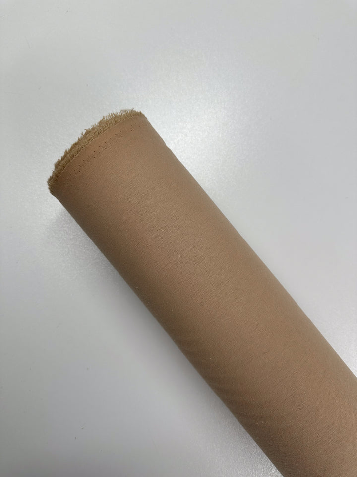 A roll of Super Cheap Fabrics' Ramie Linen in Hazelnut, 150cm wide, sits diagonally on a white surface with the edge slightly frayed at the top. The natural fiber fabric appears smooth and tightly rolled.