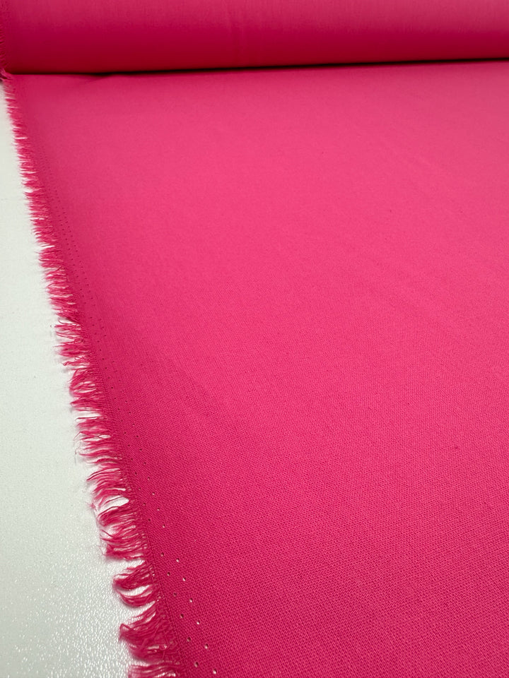 A roll of Super Cheap Fabrics' Ramie Linen in Azalea Pink, 150cm wide, laid flat. Its natural fiber showcases a smooth texture with vibrant color, and the frayed edges add depth as it stretches into the background.