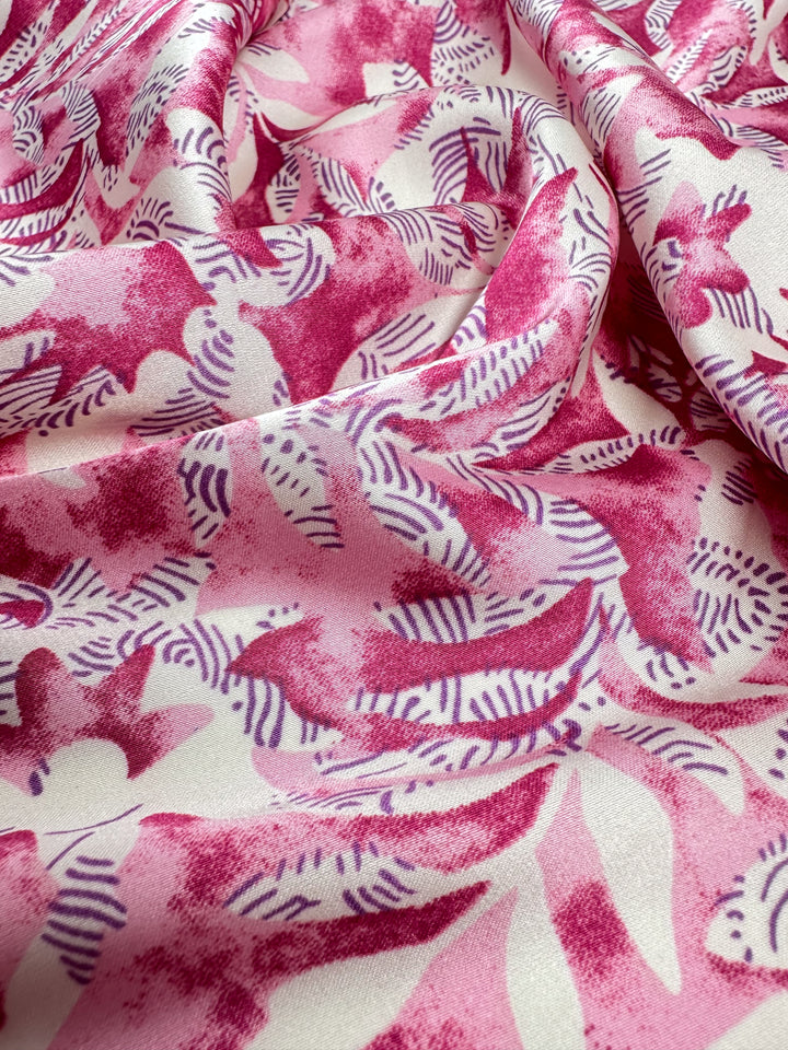 A close-up of Super Cheap Fabrics' Silky Satin - Lotus shows a lightweight satin fabric in pink and white, featuring a floral pattern with varied pink and purple shades, abstract leaf, and flower shapes, ideal for creating elegant pieces. Fabric width: 150cm.