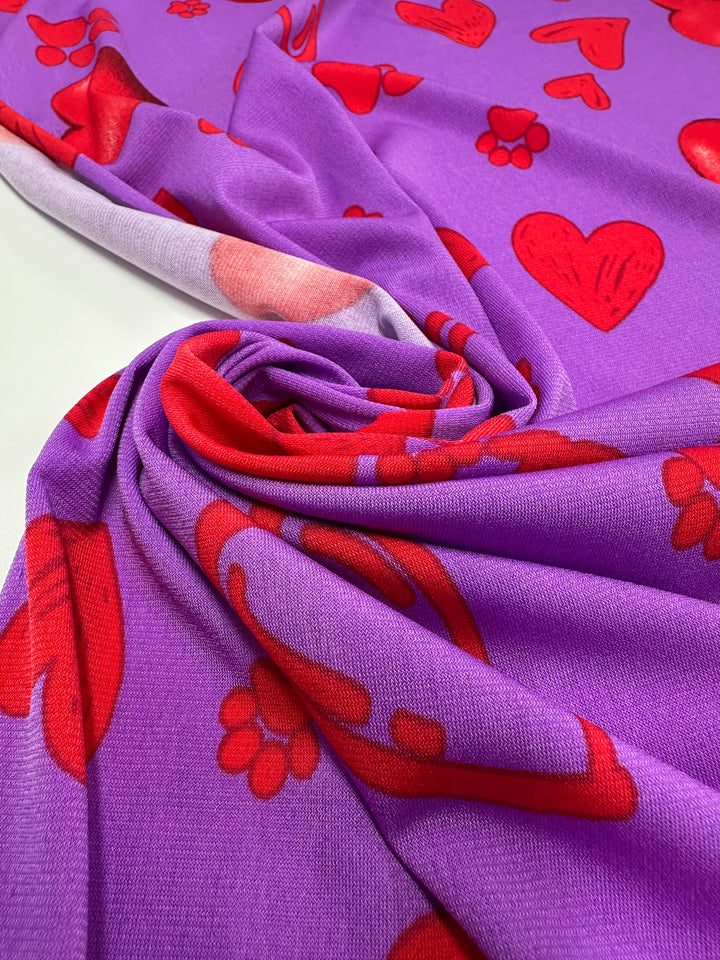A close-up of Super Cheap Fabrics' Printed Lycra - Love Riot (150cm), a medium weight fabric featuring red heart and floral patterns, gathered and spiraled on a flat surface.