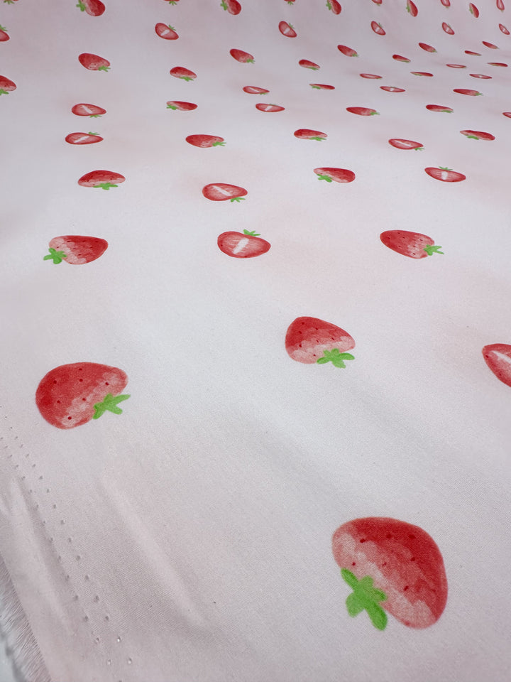 A sheet of lightweight cotton from Super Cheap Fabrics, named "Printed Cotton - Tutti Fruity - 150cm," showcases a whimsical design with a repeating pattern of small red strawberries and green leaves, crafted from natural cotton fibers.