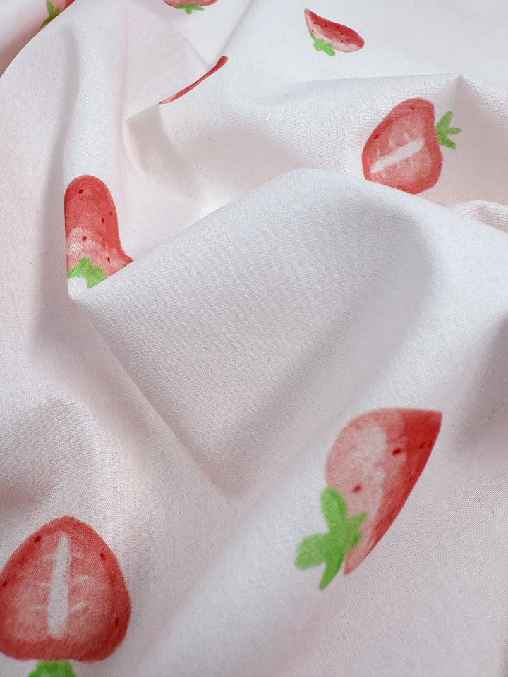 Close-up of a lightweight cotton fabric from Super Cheap Fabrics, named "Printed Cotton - Tutti Fruity - 150cm." It features a light background with a pattern of red strawberries and green leaves. The natural cotton fibers appear soft and slightly wrinkled, beautifully showcasing the repeated fruit design.