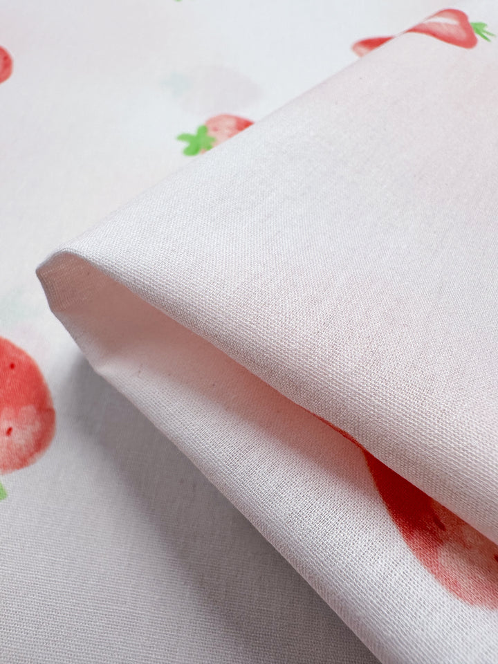 The "Printed Cotton - Tutti Fruity - 150cm" by Super Cheap Fabrics is a folded piece of white cotton fabric featuring a pattern of ripe red strawberries complemented by green leaves scattered across its surface. Crafted from natural cotton fibers, this lightweight fabric offers a smooth texture and gentle sheen, adding an elegant touch.