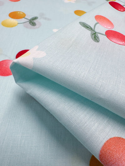 A close-up view of the Ripe Printed Cotton from Super Cheap Fabrics shows pastel blue fabric adorned with a vibrant design featuring red, yellow, and orange cherries with green leaves. The 100% cotton material, which measures 150cm in width, is neatly folded to emphasize its smooth texture and eye-catching pattern.