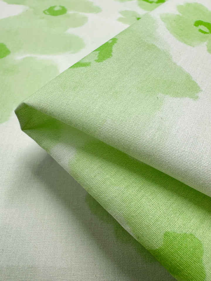 A close-up image of folded fabric featuring the Lime Bloom pattern by Super Cheap Fabrics showcases a light green floral design on a white background, crafted from Printed Cotton. This material is soft and highlights the subtle, natural texture typical of clothing and household items made with natural cotton fibers.