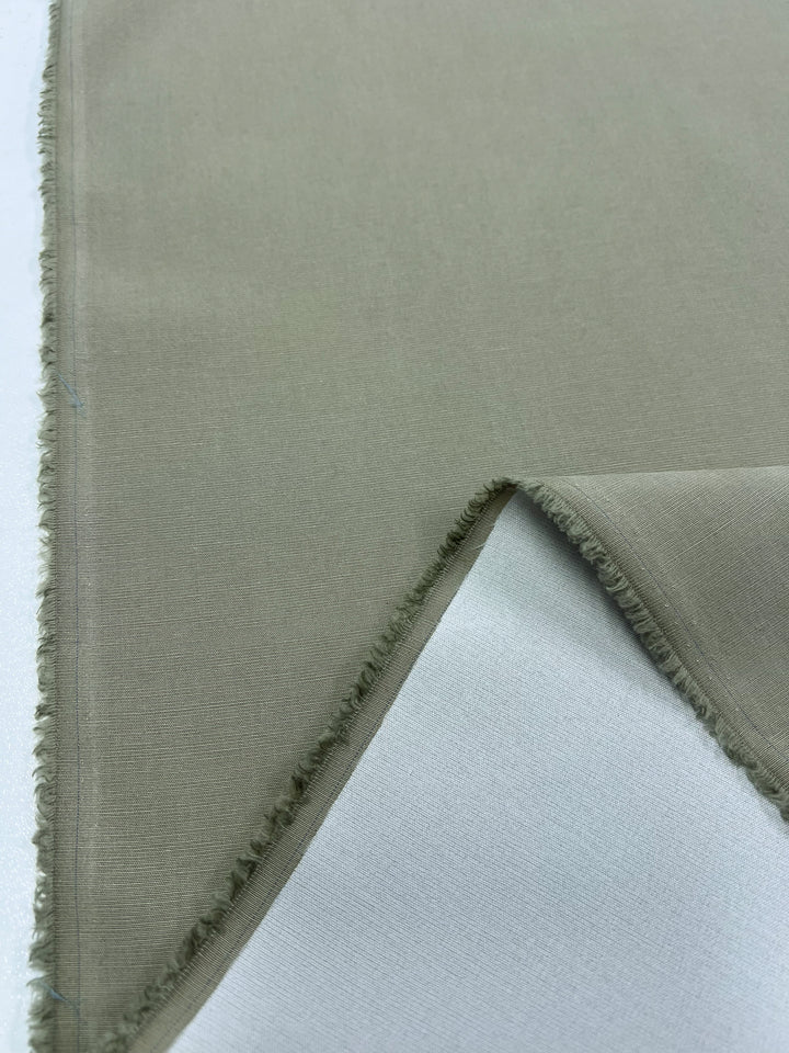 Close-up of a piece of light gray Italian Linen Blend - Elm - 150cm by Super Cheap Fabrics with frayed edges, folded back to reveal the inner side. The medium weight linen is laid out on a white surface.