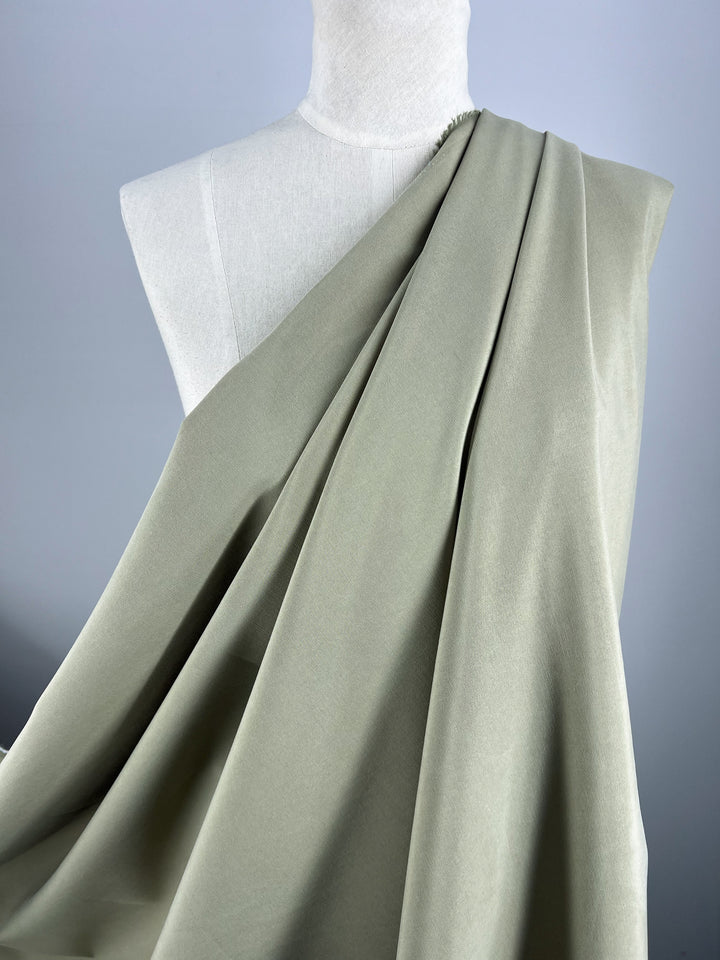 A light grayish-green Super Cheap Fabrics Linen Blend - Elm - 150cm is elegantly draped over a white mannequin on a neutral background. The cloth appears smooth and flowing, with soft folds cascading from the shoulder to the chest of the mannequin.