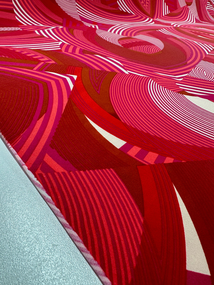 A close-up view of an abstract red and pink Printed Lycra - Wonka - 150cm from Super Cheap Fabrics, crafted from medium-weight fabric, with intricate swirling patterns and various shades of red. The design includes curved lines and geometric shapes, creating a visually engaging, dynamic texture. The fabric lays on a reflective silver floor.