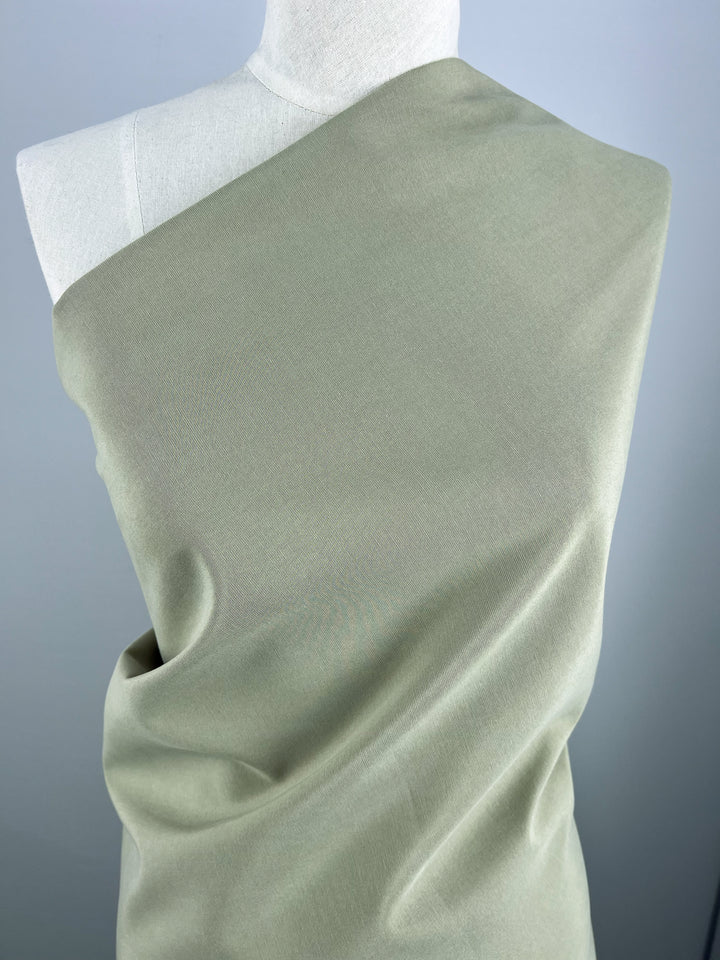 A mannequin is draped with a single piece of medium weight, light olive green Linen Blend - Elm - 150cm from Super Cheap Fabrics. The linen fabric covers the mannequin's torso diagonally, creating gentle folds and smooth contours. The background is a simple, muted grey.