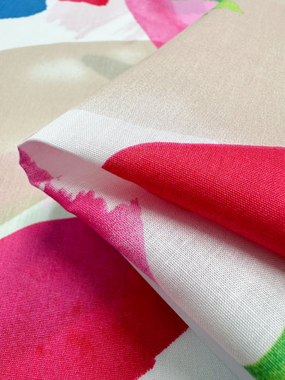 A close-up of the Super Cheap Fabrics "Printed Cotton - Salada - 150cm" showcases a folded corner, revealing its bold and abstract patterns in red, pink, beige, and green hues. This vibrant design offers a dynamic and artistic look suitable for clothing and household items.