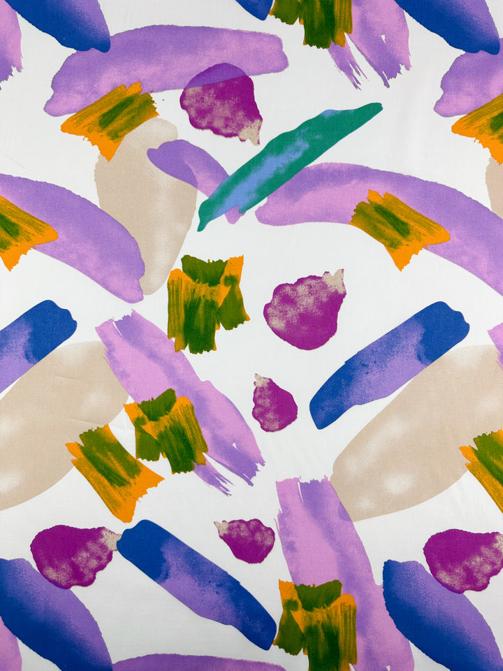 The Printed Cotton - Petals from Super Cheap Fabrics features an abstract design of brush strokes in purple, pink, green, orange, and beige on a white background. The shapes and colors are randomly dispersed across the 150cm wide fabric, resulting in a vibrant and dynamic composition.