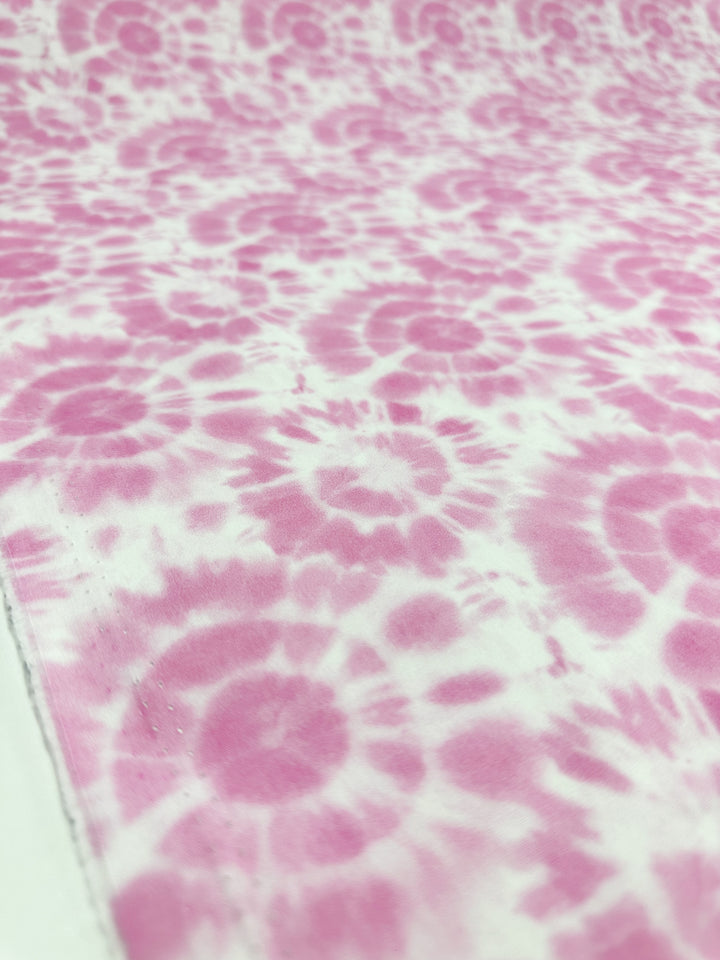 A detailed view of the Printed Cotton - Rosebloom - 150cm from Super Cheap Fabrics showcases a pattern of pink tie-dye swirls on a white cotton base. The design is both soft and vibrant, featuring repeating circular motifs crafted from natural cotton fibers, resulting in a visually dynamic texture.