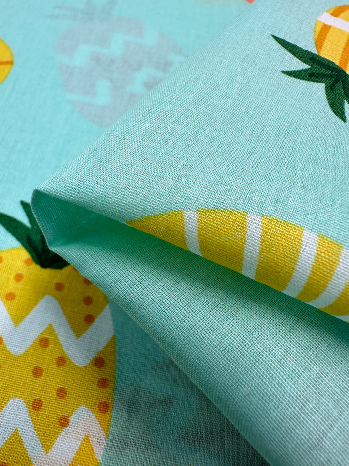 Close-up of the Super Cheap Fabrics' Printed Cotton - Pina Coalda, featuring a turquoise backdrop adorned with colorful, abstract pineapple patterns. The pineapples display zigzag and striped designs in yellow and white, accented by green leaves. Perfect for crafting clothing and household items, this 150cm wide fabric adds a vibrant touch to any project.