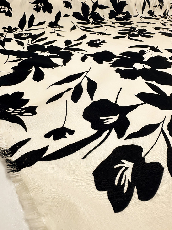 Introducing the "Printed Cotton - Visage - 150cm" by Super Cheap Fabrics, featuring a striking black floral pattern on a light beige background. The bold print of flowers and leaves creates a captivating contrast. Crafted from natural cotton fibers, the texture is evident along the edges, making it perfect for both clothing and household items.