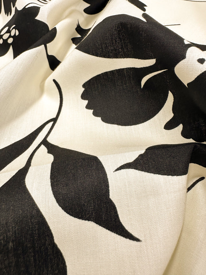 Close-up of the Super Cheap Fabrics' Printed Cotton - Visage - 150cm, showcasing a black floral pattern set against a cream backdrop. The fabric's slightly crumpled texture enhances the contrast between bold, abstract blossoms and delicate leaves, making it ideal for clothing and household items.