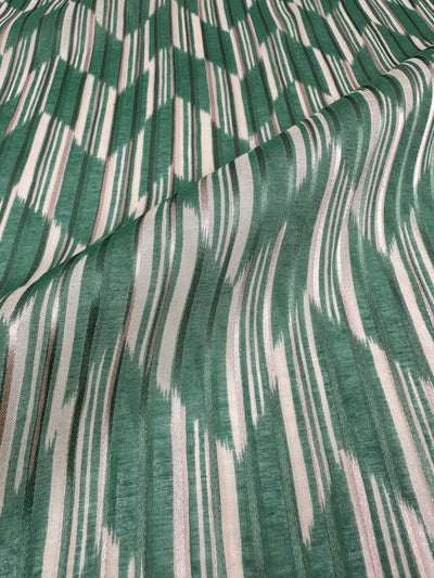 A close-up look at the Printed Silk - Escalate from Super Cheap Fabrics reveals a draping green and cream wavy striped fabric. Lightweight with visible folds, its abstract brushstroke pattern offers a textured look ideal for clothing accessories. This silk fabric spans 140cm in width.