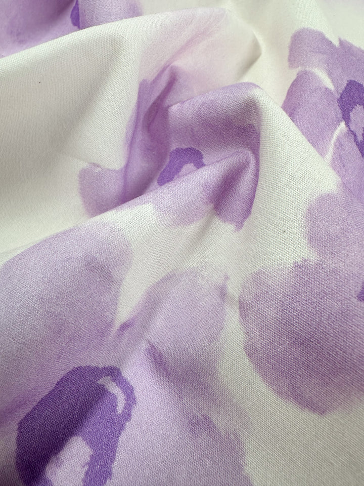 The Bellflower fabric from Super Cheap Fabrics is a soft and lightweight cotton textile, featuring abstract purple watercolor-like patterns that drape elegantly in gentle folds. The fabric is 150 cm wide and made entirely from natural cotton fibers.