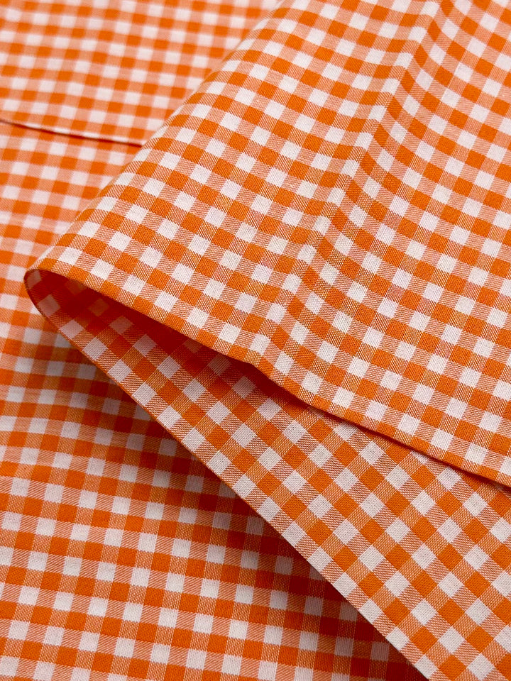 A close-up view of the Cotton Gingham Mini - Orange by Super Cheap Fabrics features a lightweight, neatly folded cotton fabric with an orange and white gingham check pattern. The natural cotton fibers display evenly spaced squares, forming a classic, vibrant design.