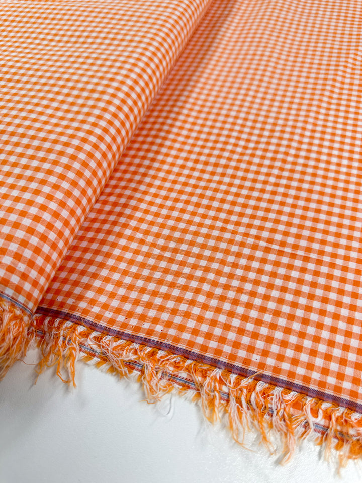 Close-up of the Cotton Gingham Mini - Orange - 112cm by Super Cheap Fabrics, showcasing its lightweight cotton fabric with an orange and white gingham pattern. The natural cotton fibers are spread flat with fringed edges, presented on a white surface.