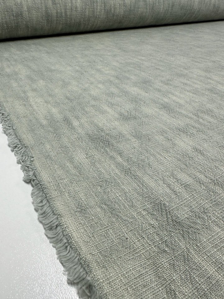 A detailed look at the Basket Dyed Cotton in Sea Mist from Super Cheap Fabrics reveals its woven texture. This heavyweight cotton fabric, featuring slightly frayed edges, is partially unrolled on a flat surface, showcasing its fine and smooth appearance. Perfect for upholstery projects, it adds elegance to any décor.