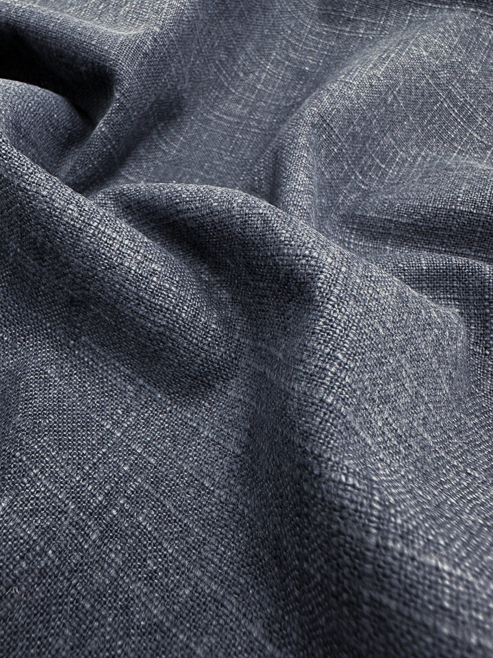 A close-up of the Basket Dyed Cotton - Navy - 140cm from Super Cheap Fabrics showcases its textured upholstery fabric with a blue-grey hue. The subtle twill weave lends it a coarse, layered appearance, while the folds in this heavyweight fabric add depth and variation in lighting.