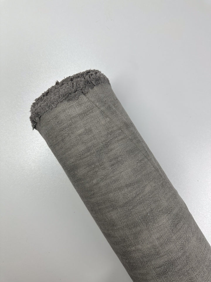 A roll of Basket Dyed Cotton in Silver Grey by Super Cheap Fabrics, noted for its textured twill weave and slightly frayed edge, is displayed on a light silver grey background.