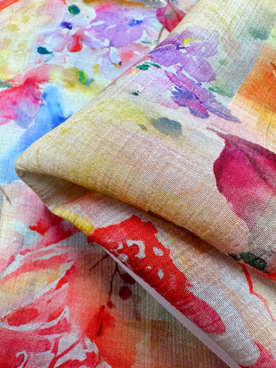 The image showcases a detailed view of the Designer Cotton - Reflection - 150cm by Super Cheap Fabrics, displaying its vibrant multicolored floral patterns in shades of red, pink, purple, and yellow on a slightly textured surface. A prominently folded section appears in the foreground, making it an ideal choice for luxury sewing projects.