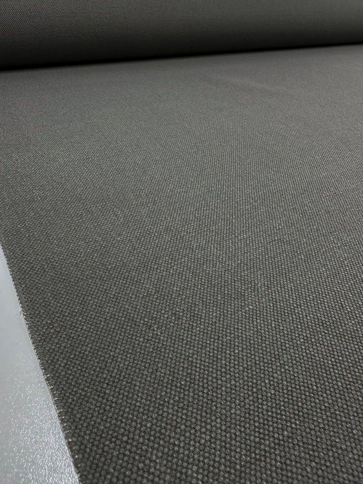 A close-up of a large roll of Canvas Linen Denim - Brushed Nickel - 140cm by Super Cheap Fabrics, unrolling on a flat surface. This dark gray upholstery fabric features a textured surface with a subtle weave pattern, offering a minimalist and industrial look ideal for modern home décor.